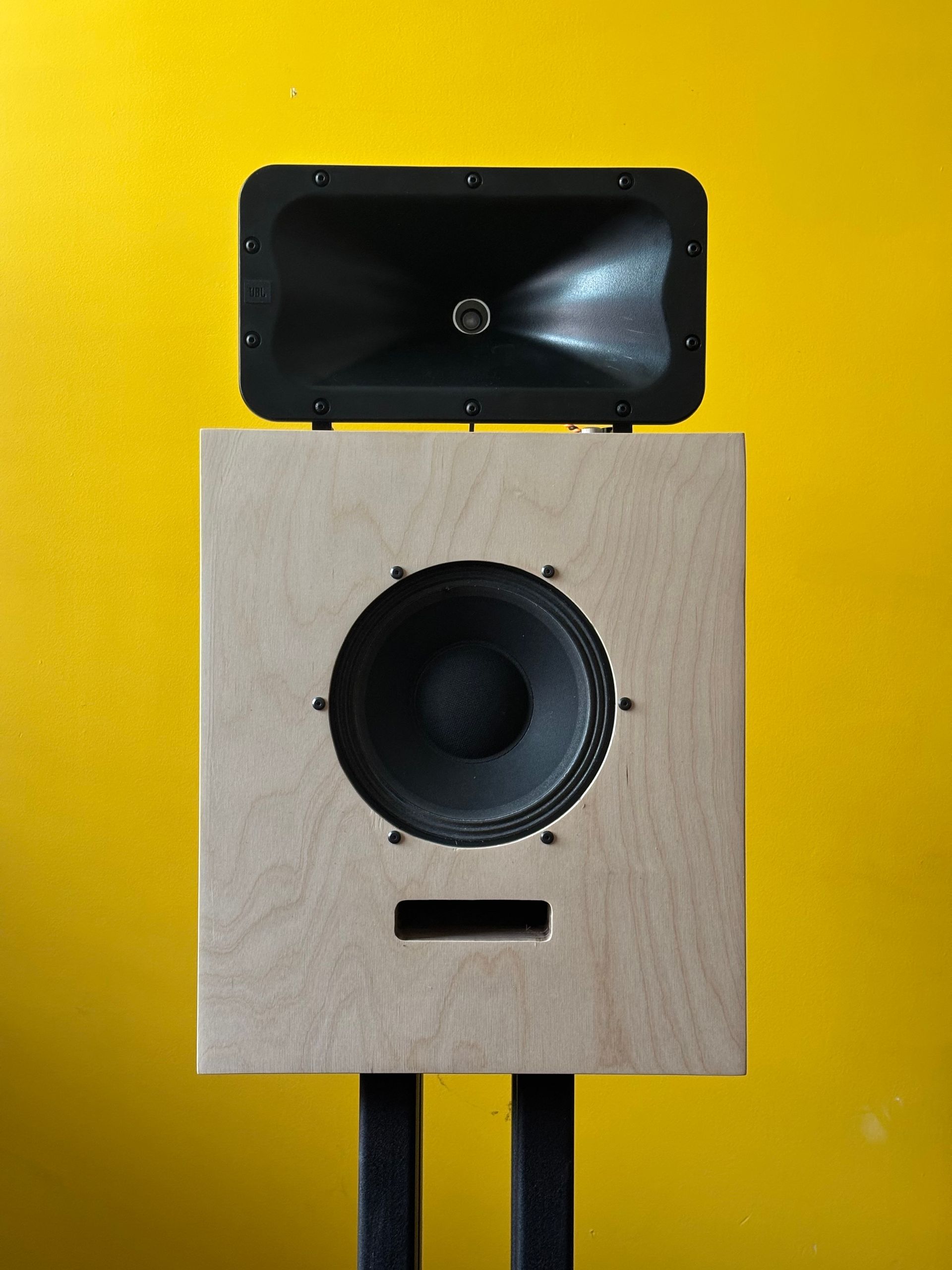 A pale birch plywood loudspeaker cabinet with a JBL horn resting on top, against a yellow wall.