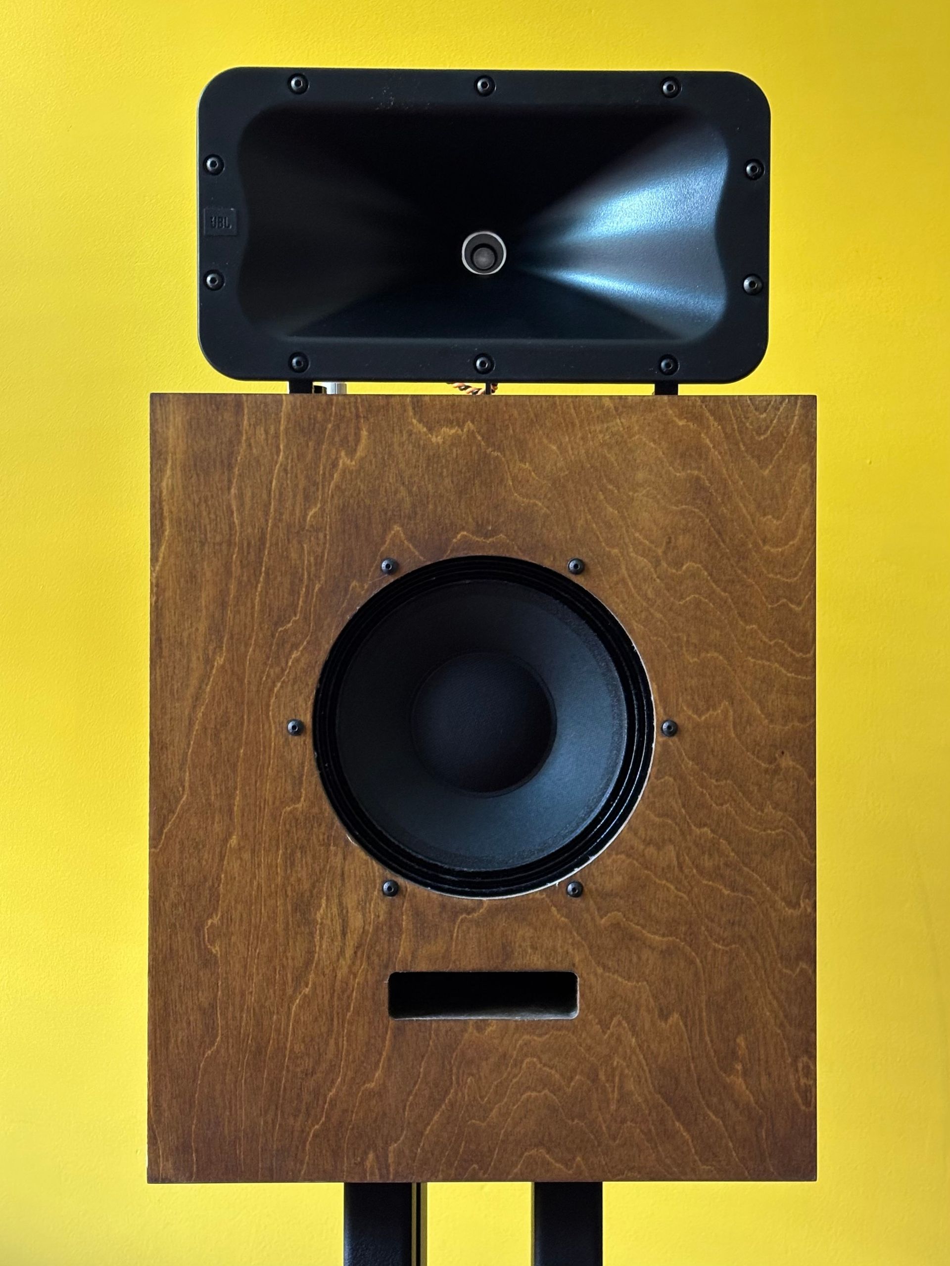 A loudspeaker cabinet in walnut-tinted birch plywood, with a black woofer, a slot port, and a JBL horn on top, against a yellow wall.