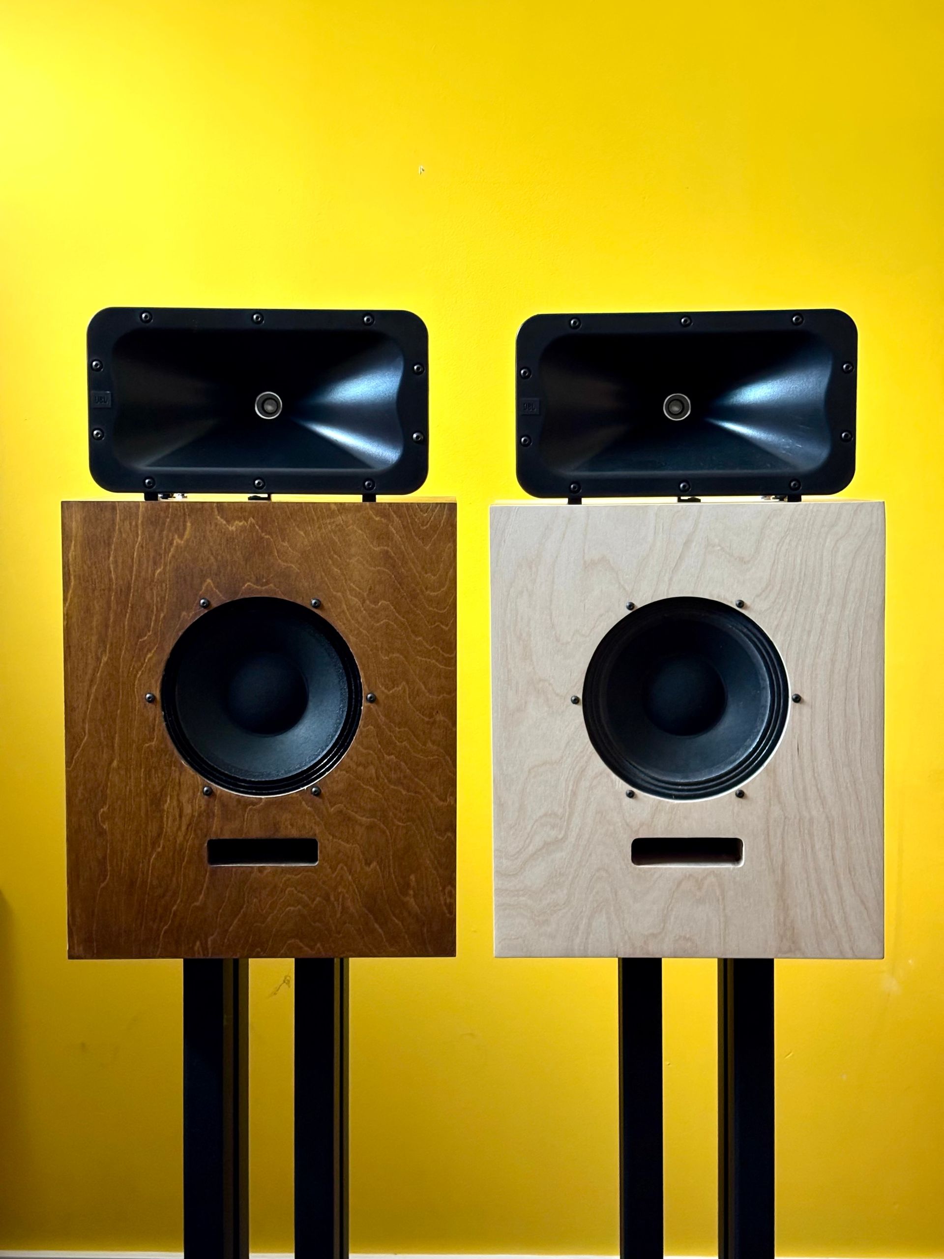 Two identical loudspeaker cabinets side by side, one in a dark walnut tint and one in pale natural birch, each topped with a JBL horn, against a yellow wall.